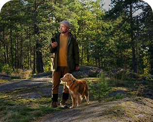 Mies ja koira metsässä. Miehellä on kädessä kännykkä.